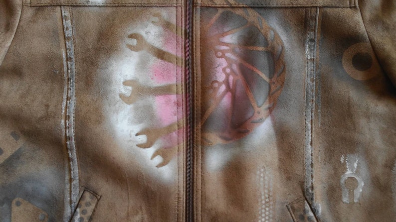 Puede incluir: Una chaqueta de ante marr&oacute;n con cierre de cremallera y un dise&ntilde;o pintado con aerosol en la parte delantera. El dise&ntilde;o presenta un patr&oacute;n abstracto rosa y blanco en el lado izquierdo y un patr&oacute;n abstracto marr&oacute;n y blanco en el lado derecho.