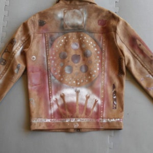 Puede incluir: Una chaqueta de cuero marr&oacute;n con un dise&ntilde;o pintado en la espalda. El dise&ntilde;o presenta un c&iacute;rculo con un patr&oacute;n de c&iacute;rculos m&aacute;s peque&ntilde;os en su interior, y un dise&ntilde;o de manos que se alzan.