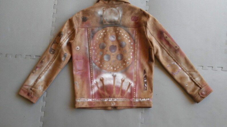 Puede incluir: Una chaqueta de cuero marr&oacute;n con un dise&ntilde;o pintado en la espalda. El dise&ntilde;o presenta un c&iacute;rculo con un patr&oacute;n de c&iacute;rculos m&aacute;s peque&ntilde;os en su interior, y un dise&ntilde;o de manos que se alzan.