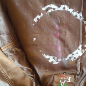 Puede incluir: El interior de una chaqueta de cuero marr&oacute;n con un c&iacute;rculo blanco con puntos alrededor. La chaqueta tiene una etiqueta que dice "EA  Authentic" en texto negro.