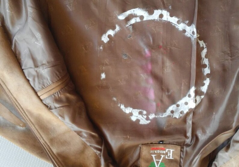 Puede incluir: El interior de una chaqueta de cuero marr&oacute;n con un c&iacute;rculo blanco con puntos alrededor. La chaqueta tiene una etiqueta que dice "EA  Authentic" en texto negro.