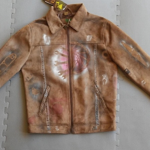 Puede incluir: Una chaqueta de ante marr&oacute;n con cierre de cremallera y un dise&ntilde;o pintado de herramientas y un coraz&oacute;n en la parte delantera. La chaqueta tiene un aspecto desgastado y est&aacute; cubierta de salpicaduras de pintura.