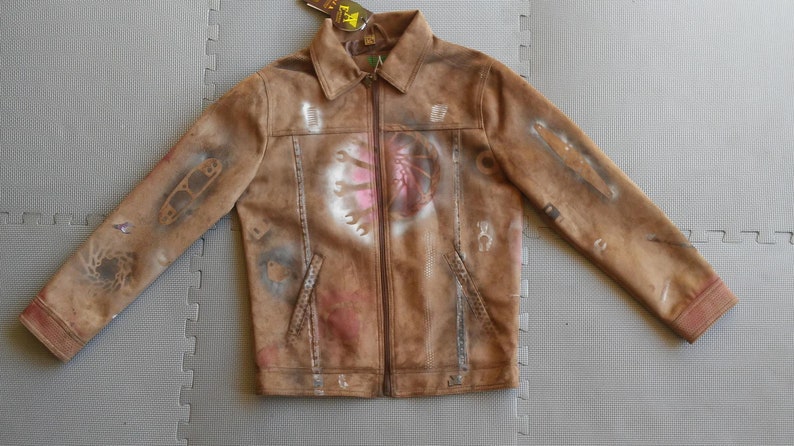 Puede incluir: Una chaqueta de ante marr&oacute;n con cierre de cremallera y un dise&ntilde;o pintado de herramientas y un coraz&oacute;n en la parte delantera. La chaqueta tiene un aspecto desgastado y est&aacute; cubierta de salpicaduras de pintura.
