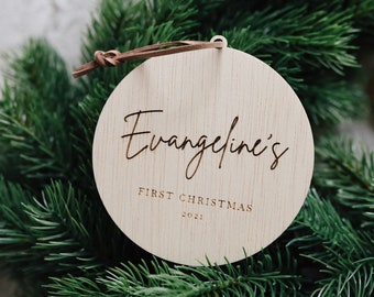May include: A round wooden ornament with the text "Evangeline's First Christmas 2021" engraved on it. The ornament is hanging from a brown leather cord and is surrounded by green pine branches.
