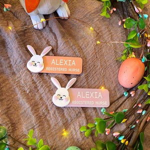 May include: Two rectangular name badges featuring a white bunny head attachment on the left side. One badge is peach and the other is light purple. Both badges display the white text ALEXIA and REGISTERED NURSE in a clean, sans-serif font.