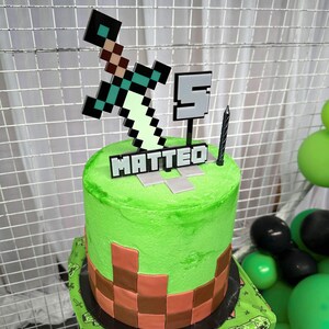 Pu&ograve; includere: Una torta di compleanno verde a tema Minecraft con una base di mattoni marroni. La torta &egrave; decorata con una spada verde, un numero 5 grigio e il nome "MATTEO" in lettere nere.