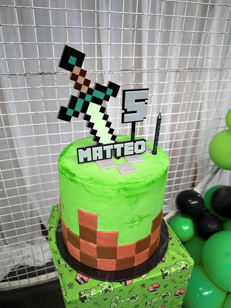 Pu&ograve; includere: Una torta di compleanno verde a tema Minecraft con una base di mattoni marroni. La torta &egrave; decorata con una spada verde, un numero 5 grigio e il nome "MATTEO" in lettere nere.