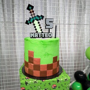Pu&ograve; includere: Una torta a due piani a tema Minecraft, verde e marrone, con un topper a forma di spada e piccone. La torta &egrave; decorata con un motivo a scacchi verde e marrone e il nome "MATTEO" &egrave; scritto in glassa bianca. La torta &egrave; su una tovaglia a tema Minecraft verde e marrone.