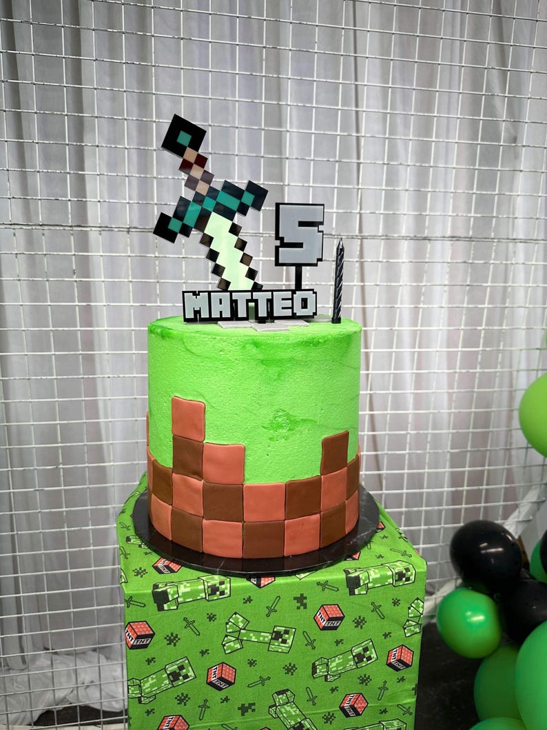 Pu&ograve; includere: Una torta a due piani a tema Minecraft, verde e marrone, con un topper a forma di spada e piccone. La torta &egrave; decorata con un motivo a scacchi verde e marrone e il nome "MATTEO" &egrave; scritto in glassa bianca. La torta &egrave; su una tovaglia a tema Minecraft verde e marrone.
