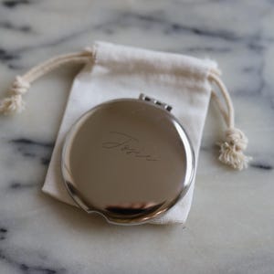 May include: A silver compact mirror with the name "Tessie" engraved on the surface, presented on a white cloth pouch. The pouch is on a marble surface. The compact is round and has a hinged opening.