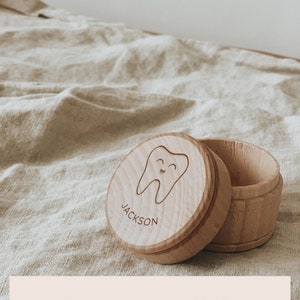 May include: A wooden tooth fairy box with a smiling tooth design and the name "JACKSON" engraved on the lid. The box is open and the lid is lying next to it. The box is on a light brown fabric surface. DESIGN C