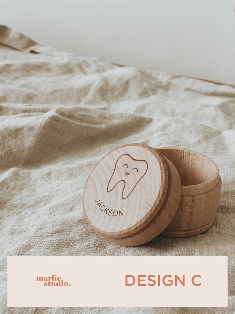 May include: A wooden tooth fairy box with a smiling tooth design and the name "JACKSON" engraved on the lid. The box is open and the lid is lying next to it. The box is on a light brown fabric surface. DESIGN C