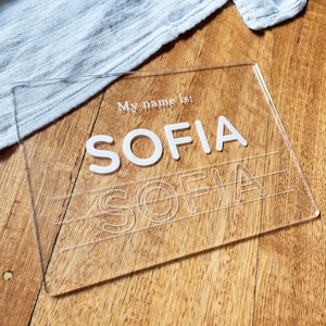 May include: A clear acrylic learning board with the text "My name is: SOFIA" printed on it. The name SOFIA is also printed in white letters on the board. There are lines underneath the name for tracing.