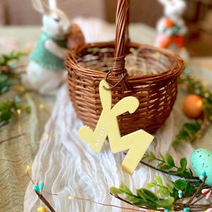 May include: A brown woven wicker basket with a single handle, featuring a pale yellow acrylic tag shaped like a rabbit ear and the letter M, with the name MATTEO engraved on the tag, hanging from the basket handle by a dark brown cord.