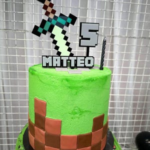 Pu&ograve; includere: Una torta a tema Minecraft verde e marrone con una spada e il numero 5 in cima. La torta &egrave; decorata con il nome "MATTEO" in lettere pixelate.