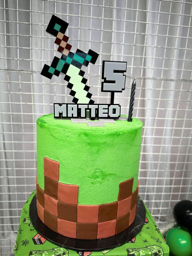 Pu&ograve; includere: Una torta a tema Minecraft verde e marrone con una spada e il numero 5 in cima. La torta &egrave; decorata con il nome "MATTEO" in lettere pixelate.