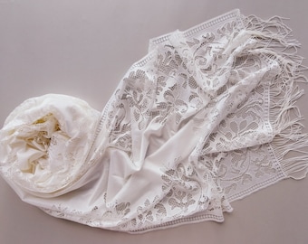 May include: A white lace scarf with a floral pattern and fringe. The scarf is draped on a light grey surface, showcasing the intricate details of the lace design. The scarf is folded and rolled up.