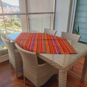 May include: A rectangular, multi-colored table runner with a striped pattern, featuring shades of red, orange, purple, and blue. The runner is placed on a glass-topped table, surrounded by four wicker chairs. The scene is set on a balcony with a city view.