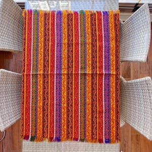 May include: A rectangular, colorful textile with vertical stripes in red, orange, yellow, purple, and green. The textile has a fringed edge and is displayed on a glass table. The design features intricate patterns within each stripe.