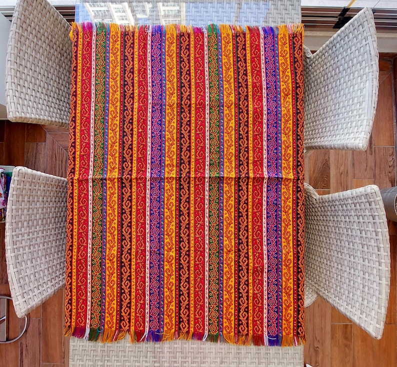 May include: A rectangular, colorful textile with vertical stripes in red, orange, yellow, purple, and green. The textile has a fringed edge and is displayed on a glass table. The design features intricate patterns within each stripe.