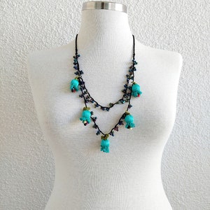 May include: A double-strand necklace with turquoise blue fabric flowers and black beads. The necklace is displayed on a white mannequin.