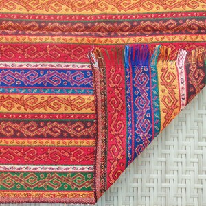 May include: A colorful woven textile with a geometric pattern. The design features horizontal stripes in red, orange, brown, blue, and green. The edge of the textile has a fringe of red, blue, and yellow threads. The textile is folded over, revealing the underside.