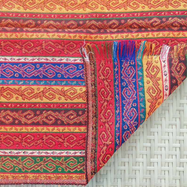 May include: A colorful woven textile with a geometric pattern. The design features horizontal stripes in red, orange, brown, blue, and green. The edge of the textile has a fringe of red, blue, and yellow threads. The textile is folded over, revealing the underside.