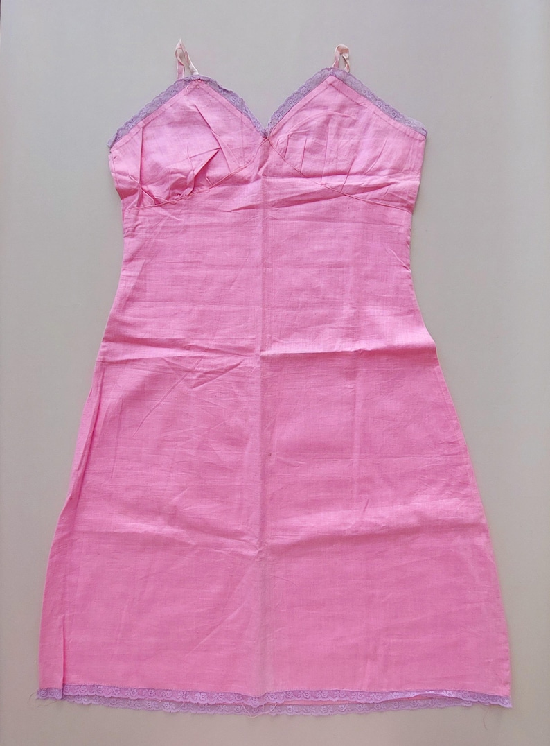 May include: A pink cotton slip dress with thin shoulder straps and delicate lavender lace trim along the neckline and hem. The dress features a fitted bodice and a slightly A-line skirt. The fabric appears lightweight and soft.  The garment is shown laid flat against a light background.
