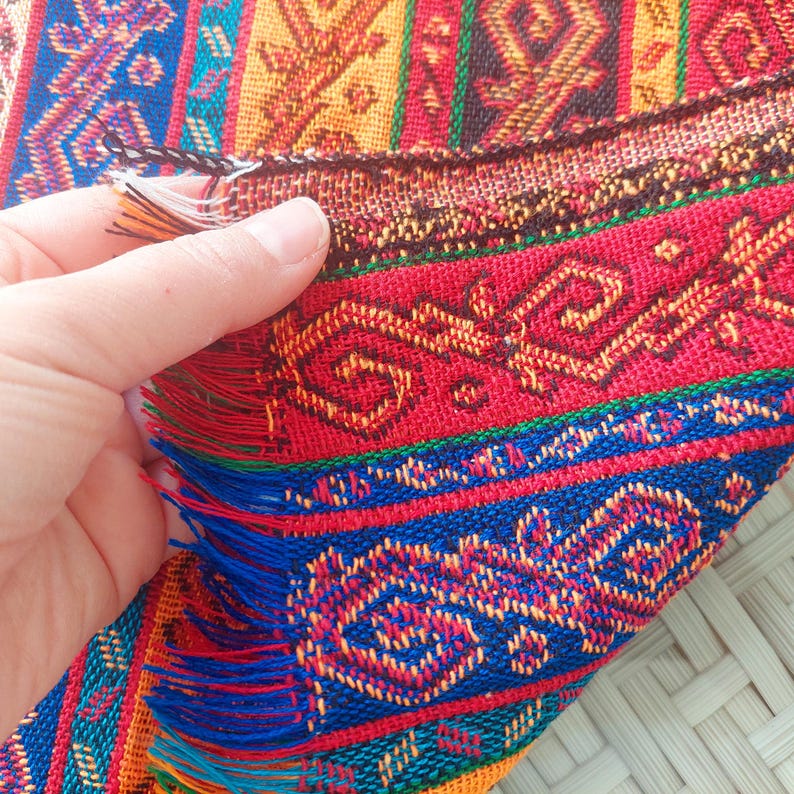 May include: Close-up of a vibrant, woven textile with intricate geometric patterns in red, blue, yellow, and green. The fabric has a fringed edge and is being held, showcasing the detailed craftsmanship and rich colors. The textile appears to be a decorative item.