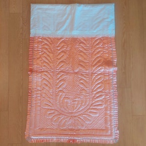 May include: A peach-colored quilted prayer rug with a white lace trim and a floral design.