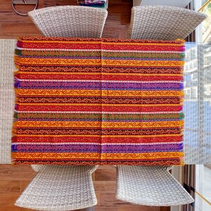 May include: A rectangular, woven textile with a vibrant, striped pattern. The design features horizontal bands of red, orange, yellow, purple, and other colors. The edges have a fringed finish. The textile is placed on a glass table, surrounded by white wicker chairs.