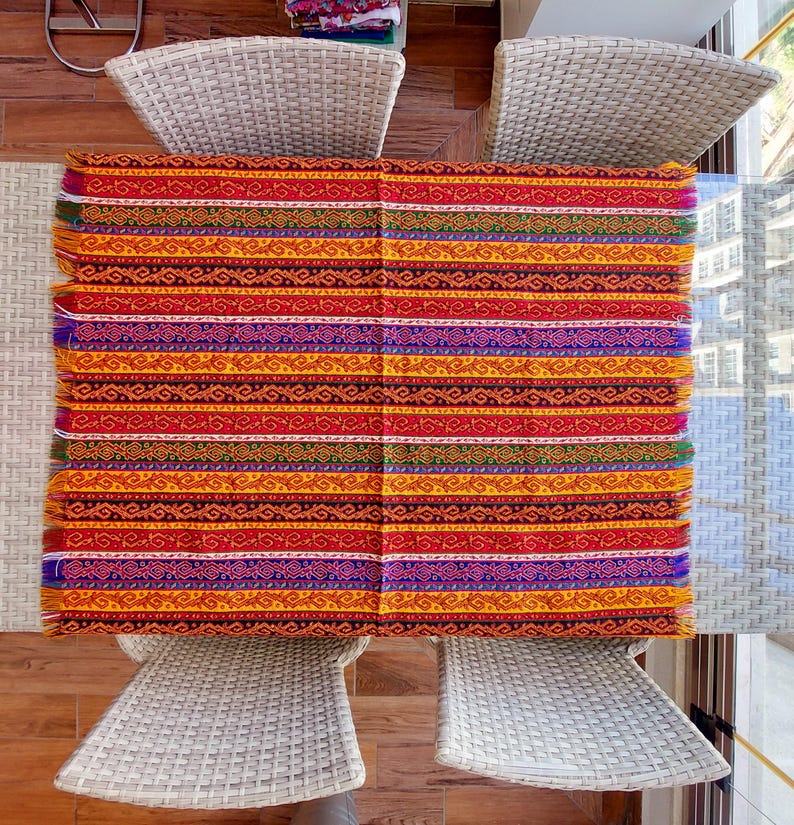 May include: A rectangular, woven textile with a vibrant, striped pattern. The design features horizontal bands of red, orange, yellow, purple, and other colors. The edges have a fringed finish. The textile is placed on a glass table, surrounded by white wicker chairs.