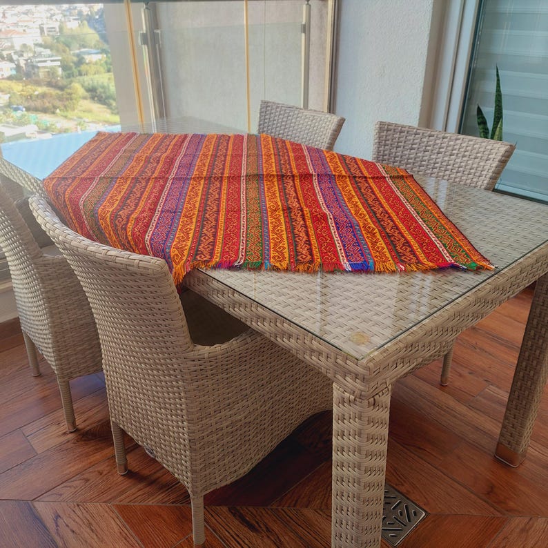 May include: A rectangular, glass-topped table with four woven wicker chairs. A colorful, striped table runner with fringed edges is draped across the table. The runner features red, orange, yellow, and purple vertical stripes.