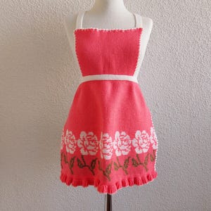 May include: A pink knitted apron with white flowers and green leaves around the bottom. The apron has white straps and a white waistband.