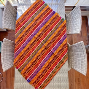 May include: A colorful, patterned table runner with a diagonal striped design. The runner features a vibrant mix of red, orange, yellow, purple, and green stripes with intricate patterns. The runner is placed on a glass table, with wicker chairs surrounding it.