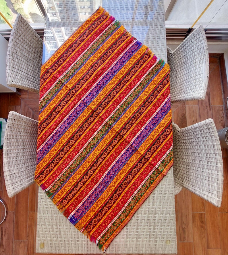 May include: A colorful, patterned table runner with a diagonal striped design. The runner features a vibrant mix of red, orange, yellow, purple, and green stripes with intricate patterns. The runner is placed on a glass table, with wicker chairs surrounding it.