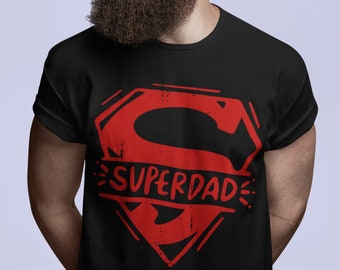 May include: Black t-shirt with a red Superman-style logo that says "SUPERDAD". The graphic is distressed, giving it a vintage look. The person in the image is wearing sunglasses and has a long beard.