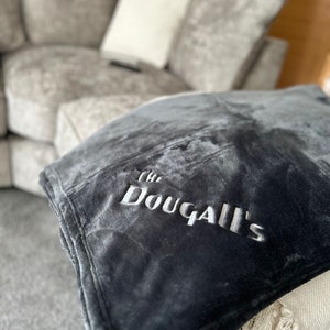 May include: A dark grey plush blanket embroidered with the white text "The Dougall's". The blanket is folded and placed on a cream throw with fringing. A grey sectional sofa is visible in the background.