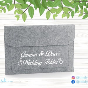 May include: A grey felt wedding folder with a flap. The folder is inscribed with "Gemma & Dave's Wedding Folder" in white script, featuring a ring graphic. The background shows green leaves and a white wood-grain pattern.