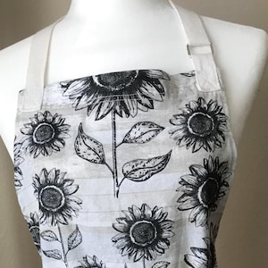 May include: A white apron with a black and white sunflower pattern. The apron has a white neck strap and waist ties. The sunflowers are detailed with leaves and stems. The apron is displayed on a mannequin.