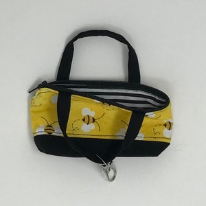 Pode incluir: Pequena bolsa tote amarela com um padrão de abelhas e detalhes em preto. A bolsa tem alças pretas e fecho de zíper. O forro interior apresenta listras pretas e brancas. Um fecho prateado está preso na parte inferior.