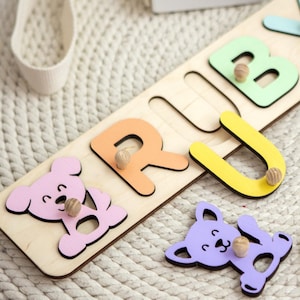 May include: Wooden puzzle spelling "RUBY" with colorful letters and wooden pegs. The letters are orange, green, yellow, and blue. Includes pink and purple bear-shaped puzzle pieces. White crocheted hats are in the background.