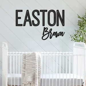 May include: Black wooden name sign that reads "EASTON Brown" in a nursery setting. The sign is mounted on a white shiplap wall above a white crib with a gray and white blanket. A potted plant is to the right.