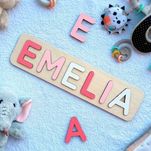 May include: Wooden name puzzle spelling "EMELIA" in pink and white letters. The puzzle is on a light wood base. Baby toys, including a stuffed elephant and a bear, surround the puzzle on a white surface.