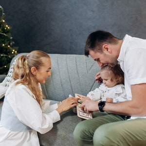 May include: A family of three playing a wooden puzzle game on a gray couch. The puzzle has colorful shapes and a light wood frame. The family is smiling and having fun together.