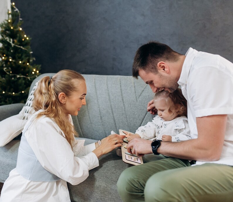 May include: A family of three playing a wooden puzzle game on a gray couch. The puzzle has colorful shapes and a light wood frame. The family is smiling and having fun together.