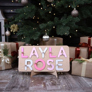 May include: A rectangular wooden name puzzle board featuring the name LAYLA ROSE in alternating pink and white letters. Each letter is a separate piece with a small wooden knob for grasping, set against a light-coloured wooden base with a stand.