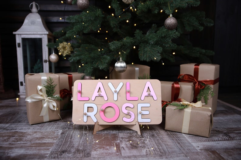May include: A rectangular wooden name puzzle board featuring the name LAYLA ROSE in alternating pink and white letters. Each letter is a separate piece with a small wooden knob for grasping, set against a light-coloured wooden base with a stand.
