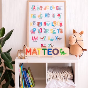 May include: A wooden name puzzle spelling MATTEO in colourful letters with a green dinosaur icon, placed in front of a framed alphabet poster featuring animal illustrations for each letter, set on a white shelf with books and a plush horse toy.
