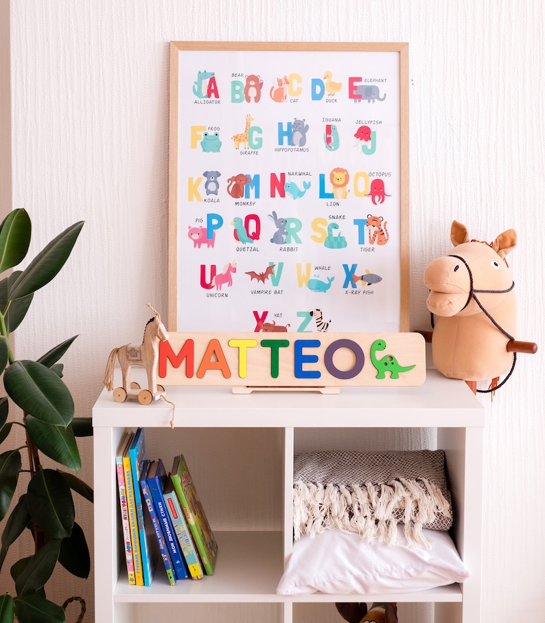 May include: A wooden name puzzle spelling MATTEO in colourful letters with a green dinosaur icon, placed in front of a framed alphabet poster featuring animal illustrations for each letter, set on a white shelf with books and a plush horse toy.